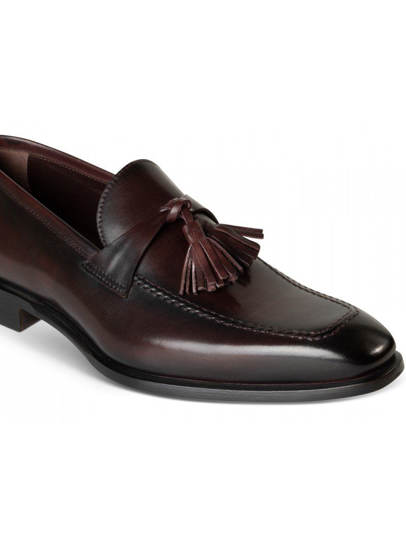 Bordeaux leather patina moccasin shoe seen from the front with two burgundy tassel fringes, burgundy ribbon, and black sole.